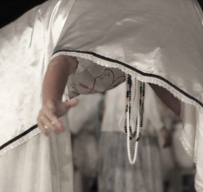 Fotografía artística persona en trance con vestimenta blanca inclinada durante ritual espiritual, collares ceremoniales colgando, fondo oscuro