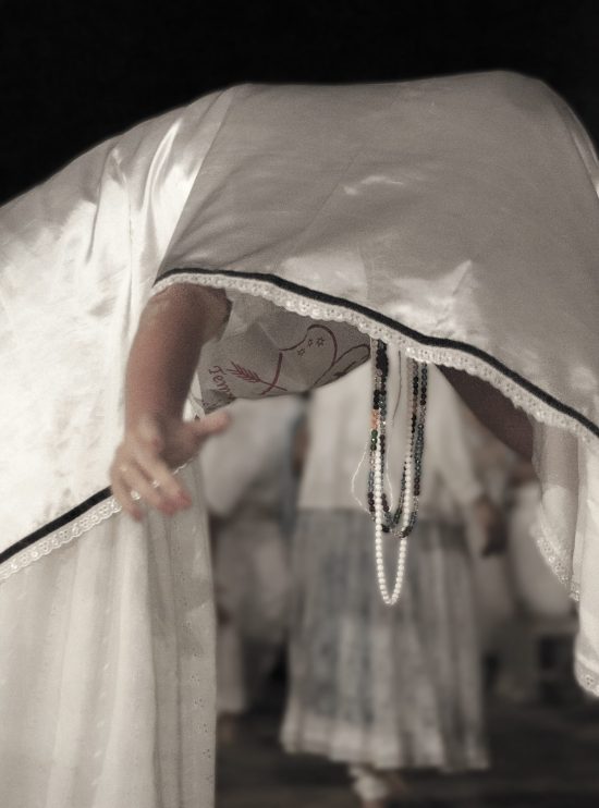 Fotografía artística persona en trance con vestimenta blanca inclinada durante ritual espiritual, collares ceremoniales colgando, fondo oscuro