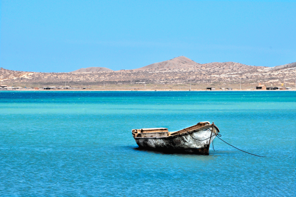 Fotografía artística de una lancha de pescador vieja en aguas turquesas del mar Caribe en La Guajira, Colombia, con un paisaje costero árido al fondo.