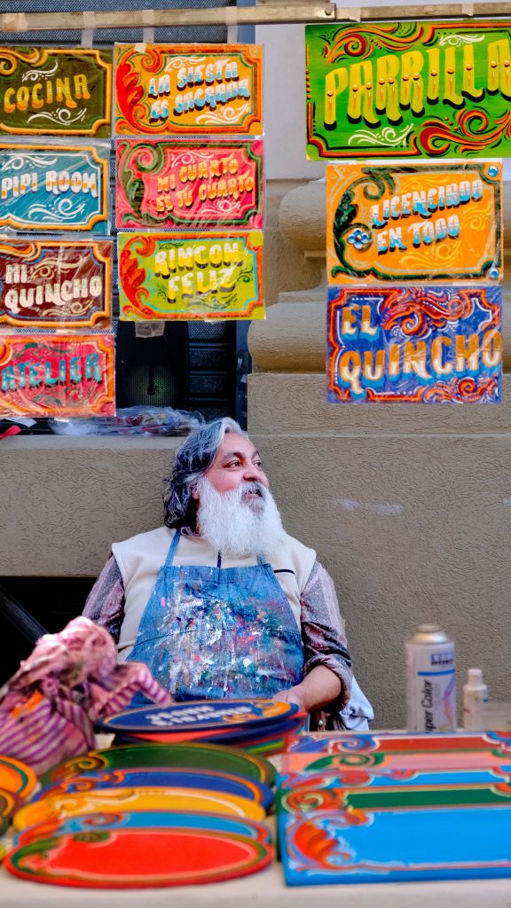 Fotografía artista fileteador porteño, barba blanca, delantal pintura, exhibiendo carteles coloridos fileteado, arte popular argentino