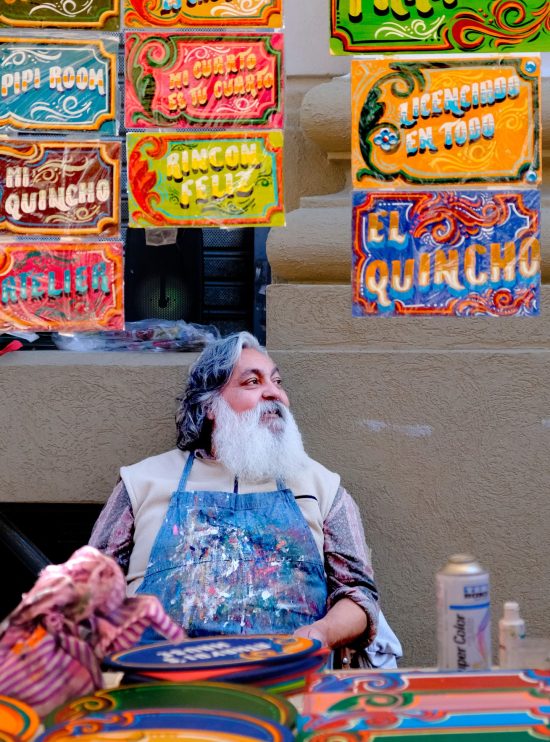 Fotografía artista fileteador porteño, barba blanca, delantal pintura, exhibiendo carteles coloridos fileteado, arte popular argentino