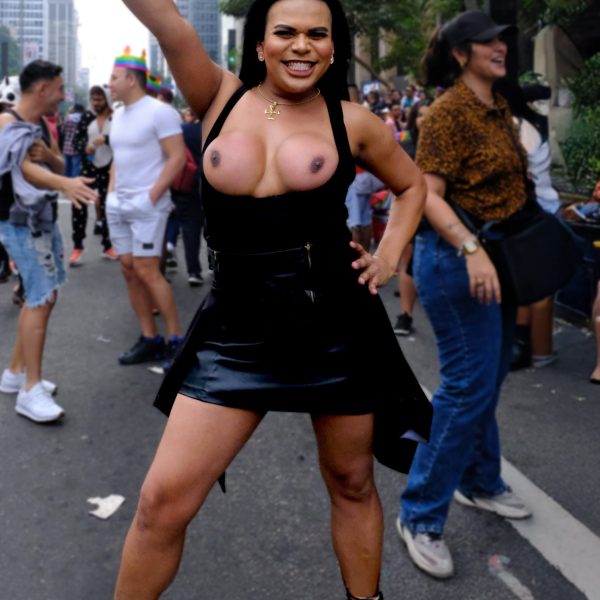 Fotografía persona trans sonriendo con vestido negro y botas en Desfile del Orgullo LGBTQ+ Sao Paulo, multitud y edificios al fondo