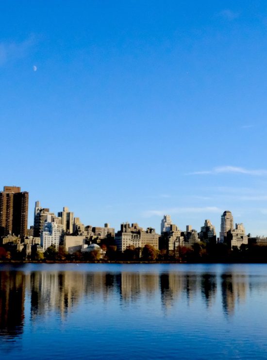 Fotografía panorámica del skyline de Nueva York reflejado en el lago de Central Park, con edificios de Manhattan y un cielo azul claro.