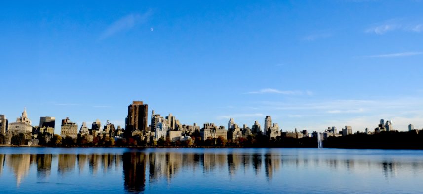 Fotografía panorámica del skyline de Nueva York reflejado en el lago de Central Park, con edificios de Manhattan y un cielo azul claro.