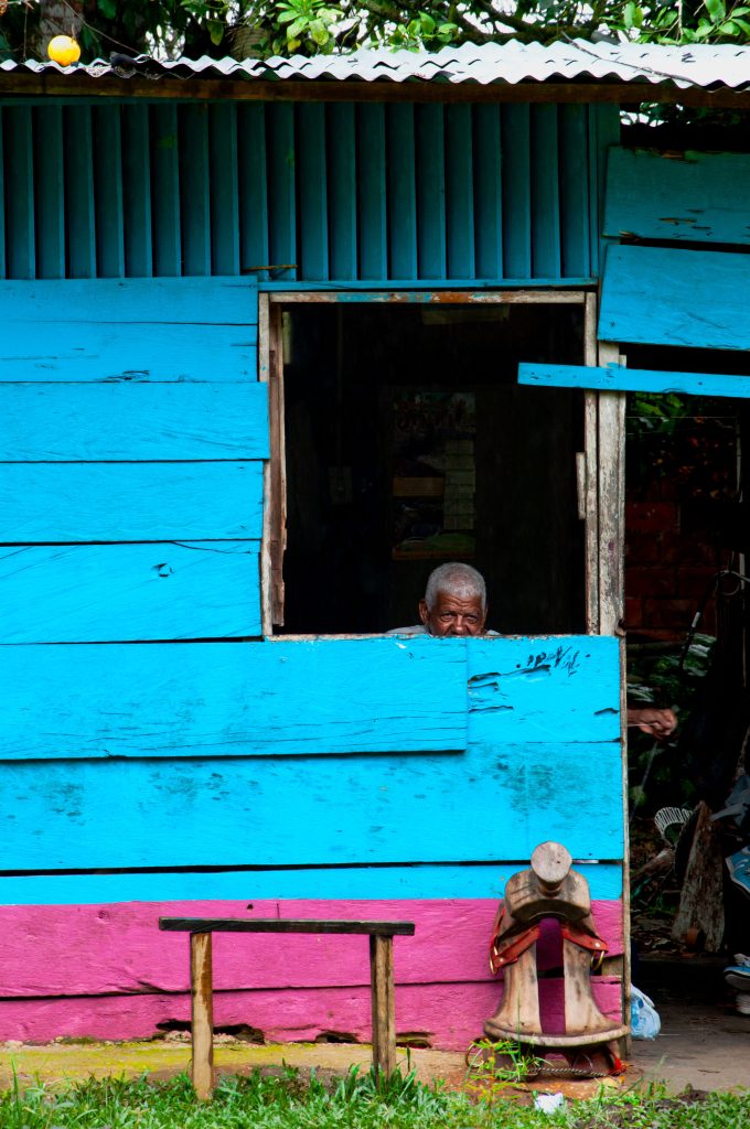 Fotografía artística hombre anciano tejedor asomado ventana casa madera azul y rosa, silla de montar exterior, vida rural