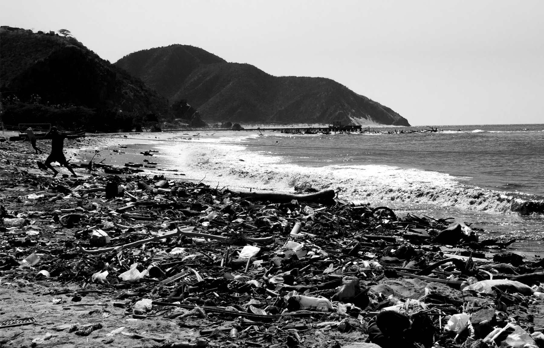 Fotografía blanco y negro de playa contaminada con basura y un pescador al fondo.